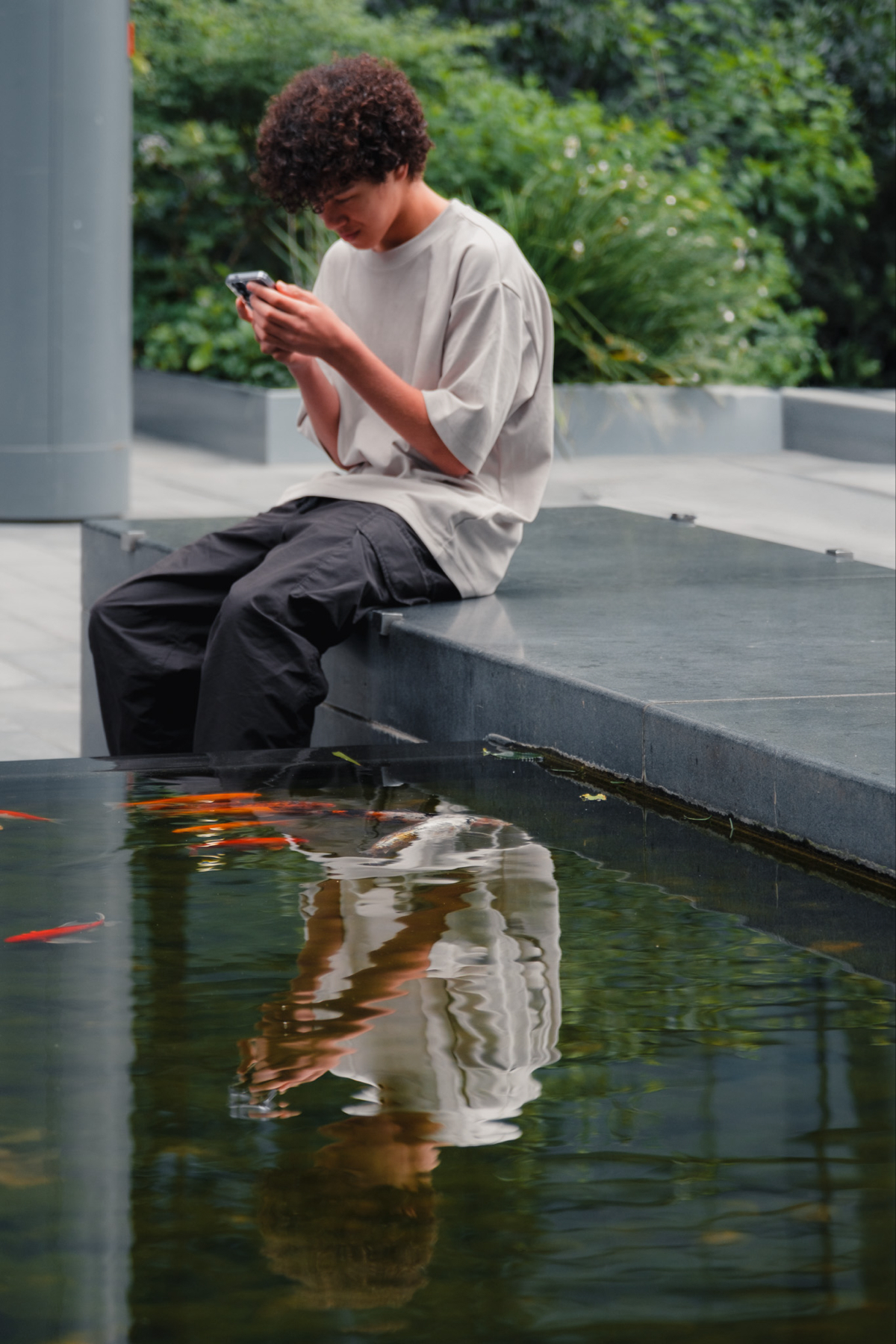 A young person with curly hair sits on the edge of a concrete ledge, focused on their phone. They are wearing a loose-fitting beige T-shirt and black pants. Below them is a pond with several orange fish swimming, and the person's reflection is