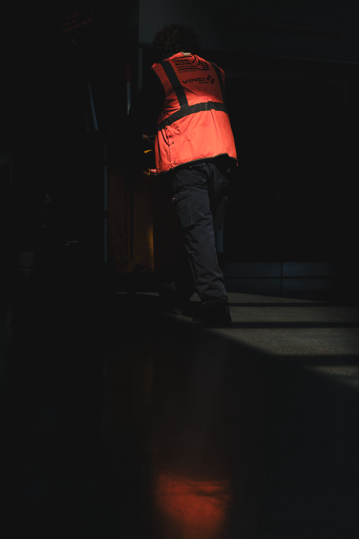 Auto-generated description: A person wearing a reflective orange vest is pushing a cart in a dimly lit environment.