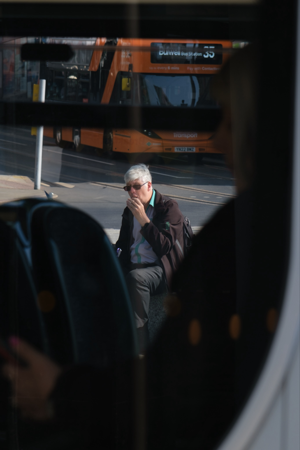 A man with gray hair, wearing sunglasses, is sitting at a bus stop with an orange bus in the background. He appears to be eating something and is dressed in a dark jacket with a light-colored shirt. The image is taken through a window,