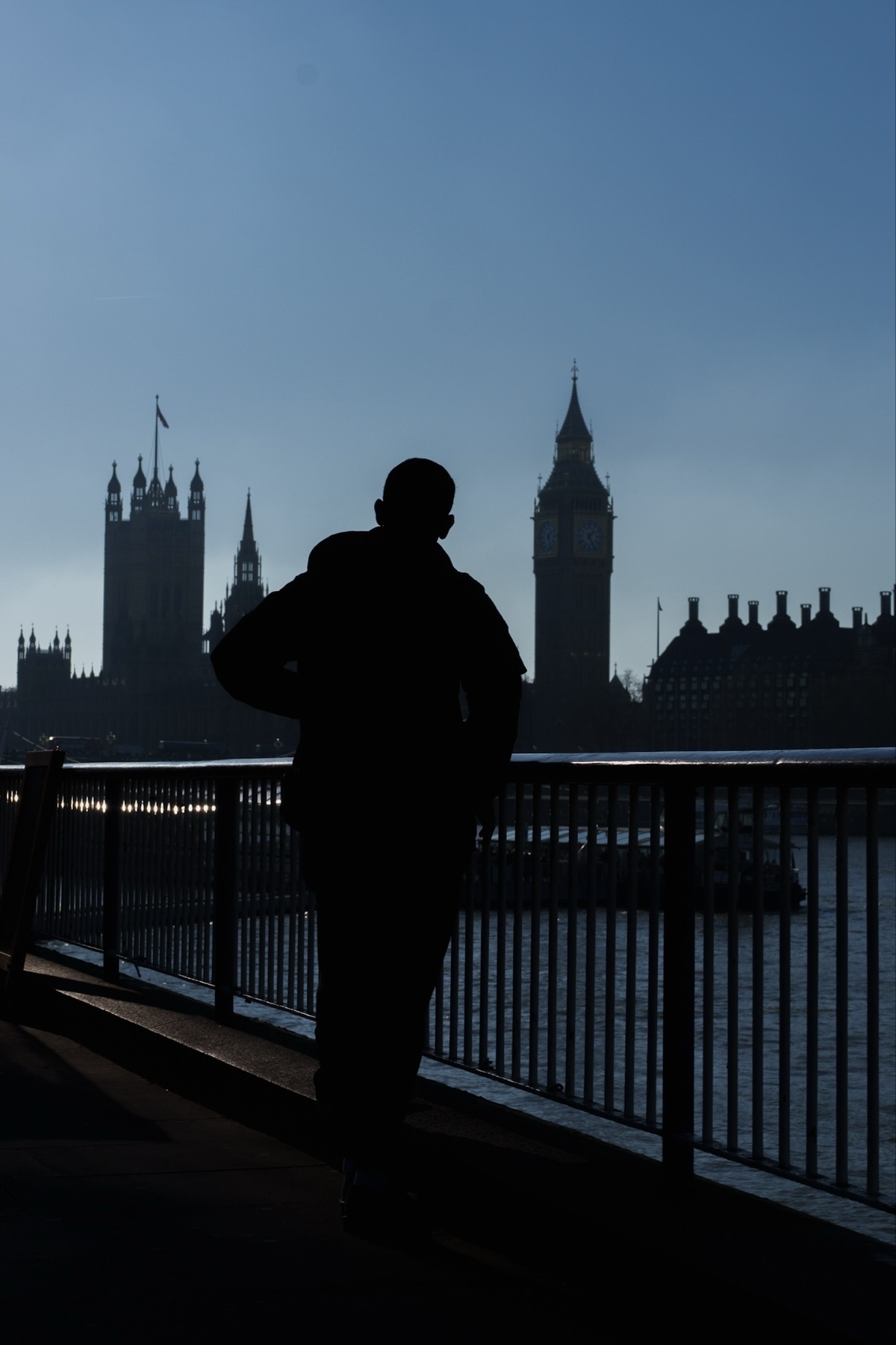 Silhouette of a person standing near a railing by the River Thames, with the Houses of Parliament and Big Ben visible in the background under a clear blue sky.