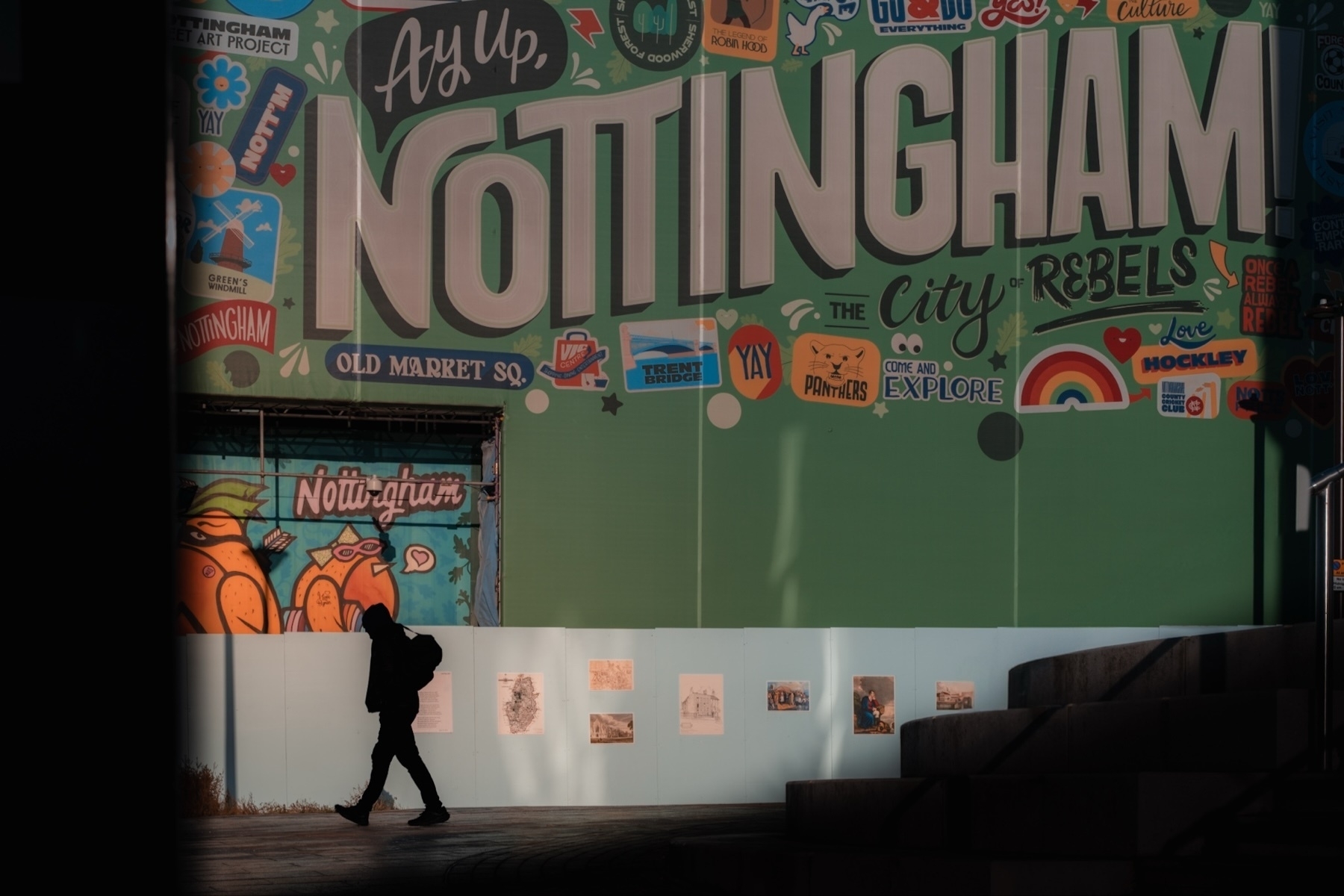 A silhouette of a person walking past a colorful mural in Nottingham. The mural features large text reading "Ay Up, Nottingham" and includes various local references, graphics, and phrases. The background is primarily green, with illustrations and vibrant details.