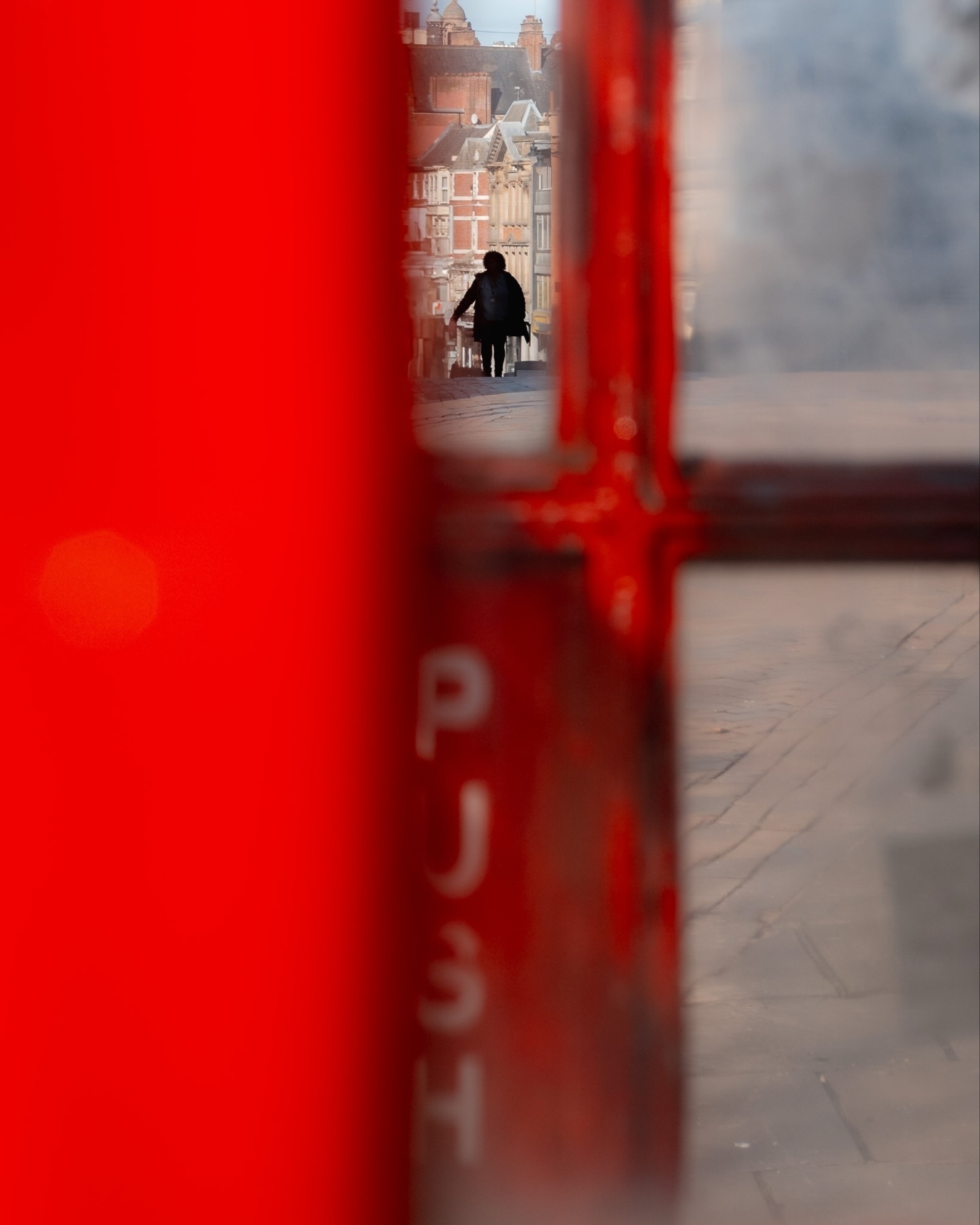An abstract view from inside a red British phone booth, focusing through its glass window onto a silhouette of a person walking in the distance on a street lined with old brick buildings. The word "PUSH" is partially visible on the glass.