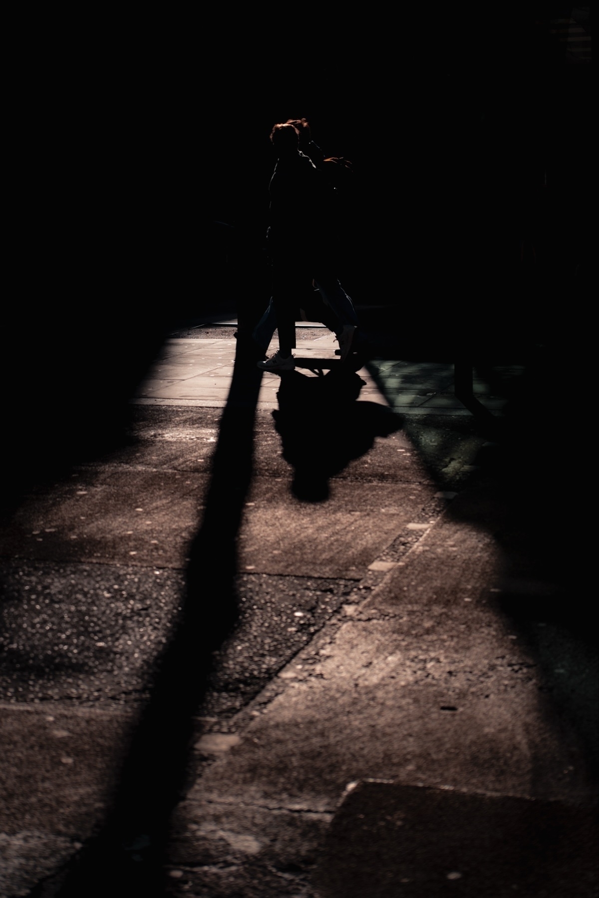 A silhouette of a person walking on a dimly lit street, casting a long shadow on the pavement. The surrounding area is mostly in darkness.
