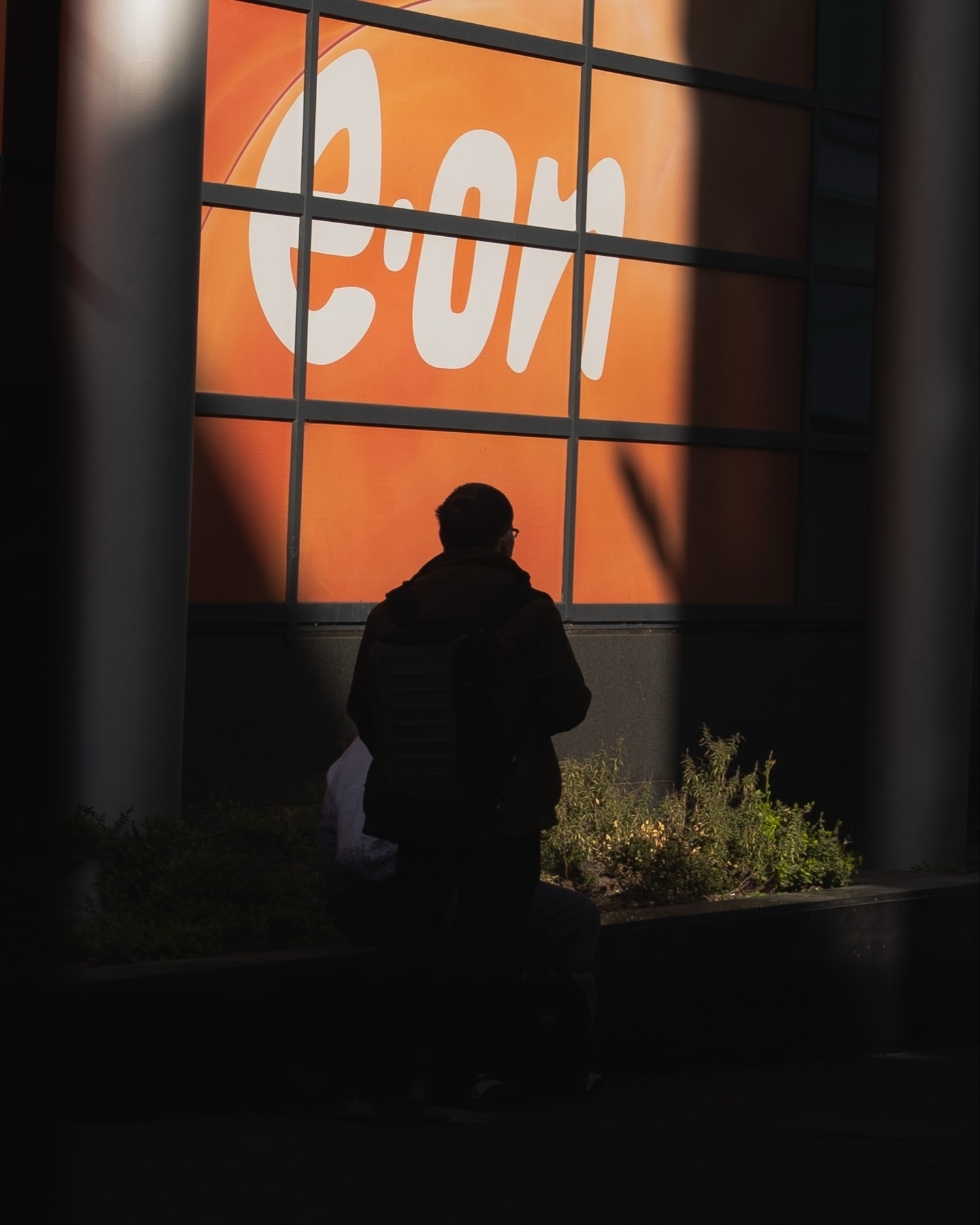 A person with a backpack stands in shadow in front of a large orange sign featuring the "E.ON" logo.
