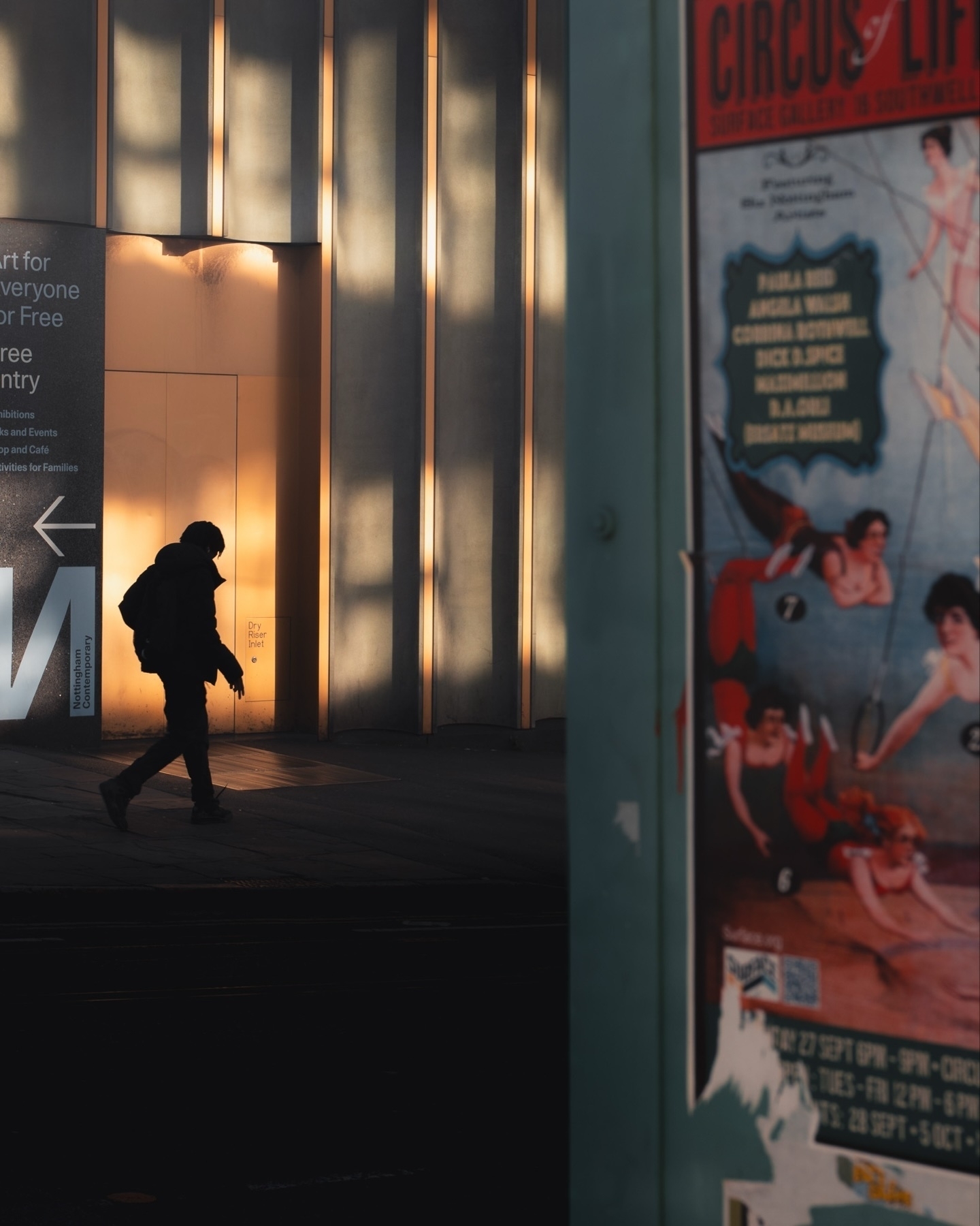 A silhouetted person walks past a modern building with vertical light panels creating shadows. A poster for a circus event with vintage-style performers is visible on the right. The area appears to be an urban setting.