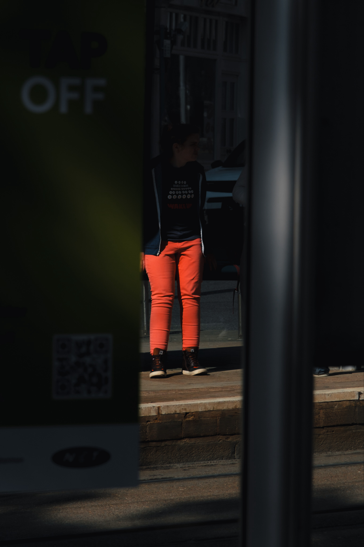 A person wearing bright orange pants and dark shoes stands partially obscured by dark shadows and a green sign with white text that reads TAP OFF. The background includes a sidewalk and part of a vehicle.