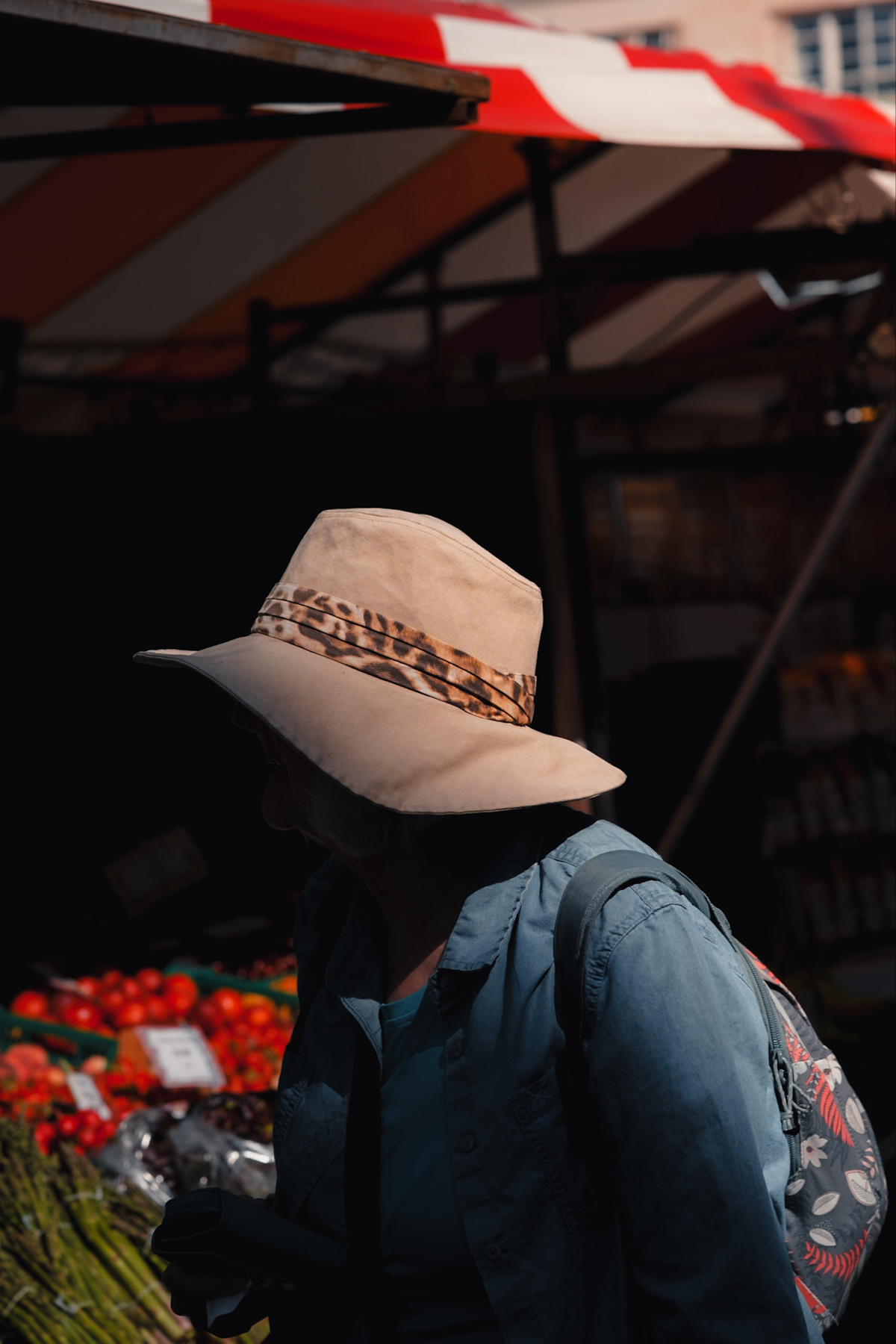 A person wearing a beige hat with a leopard print band and a blue shirt is seen in silhouette against a market stall background. There are fresh produce items such as asparagus and tomatoes visible in the background. The person has a backpack with a floral pattern.
