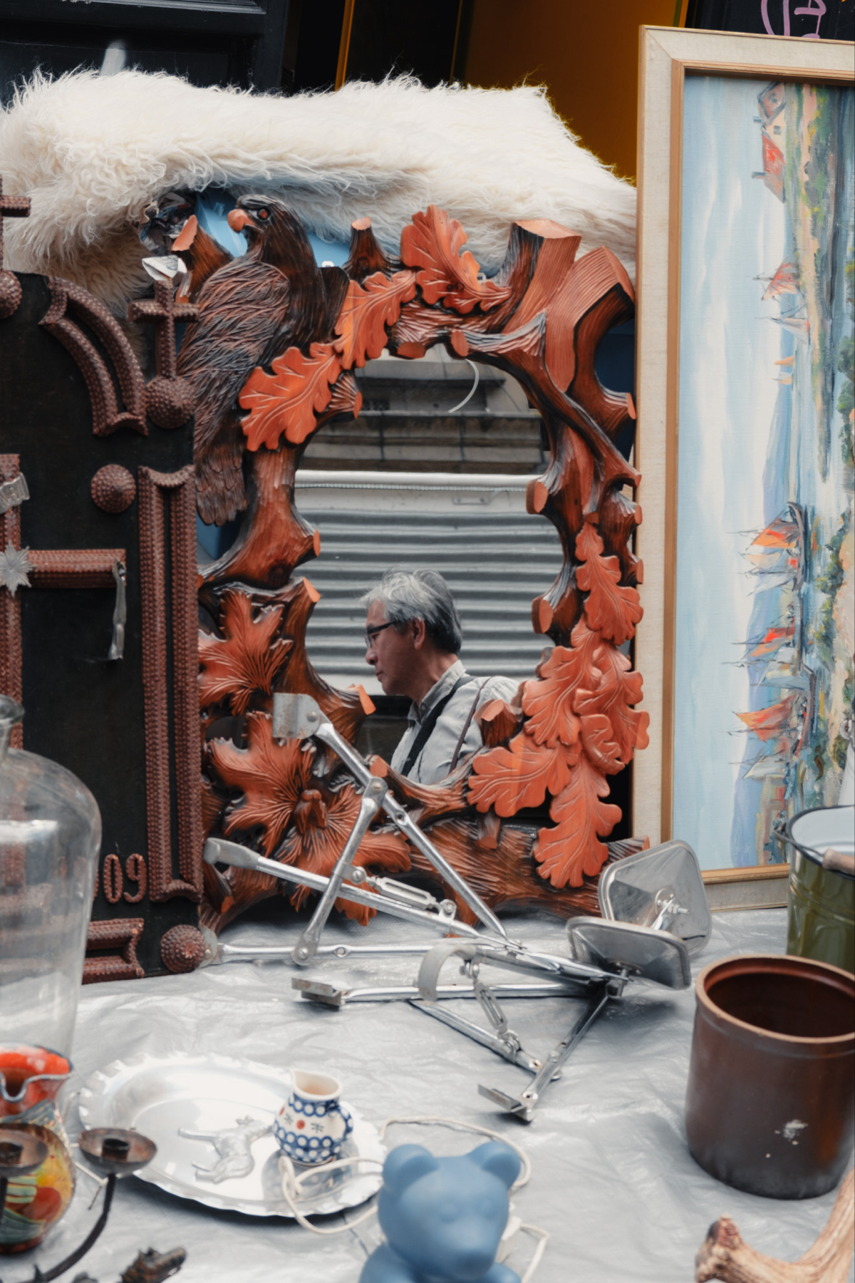 A variety of items displayed at a flea market, including an ornate mirror with a carved wooden frame featuring birds and leaves, a framed painting of colorful sailboats, and various vintage tools and trinkets. The mirror reflects an older man standing nearby.