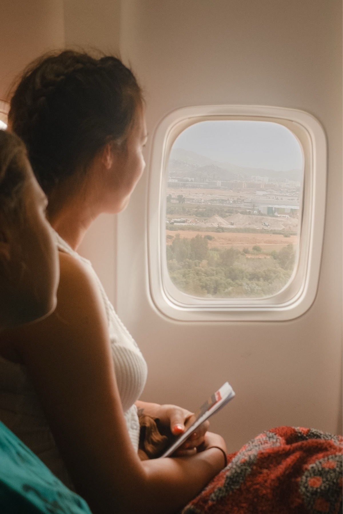 A person is looking out of an airplane window holding a book, with a view of the landscape below.