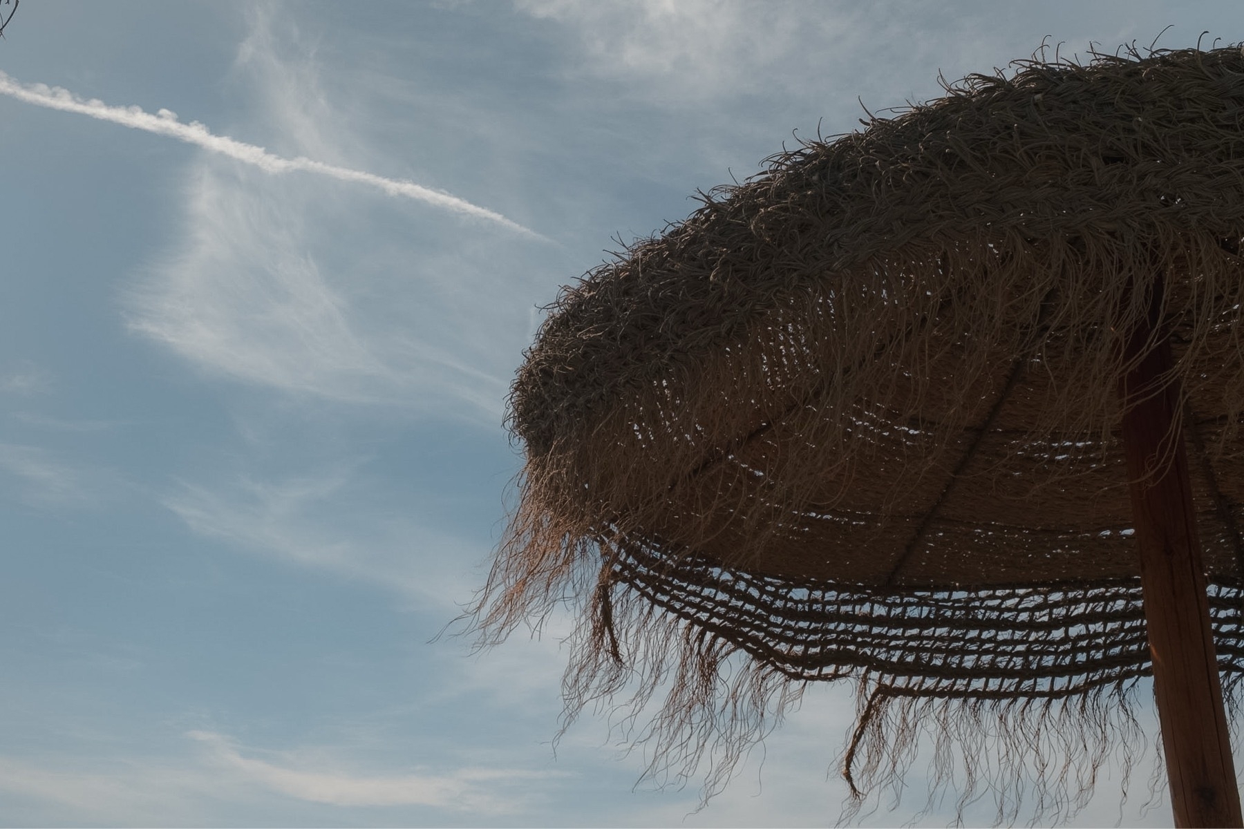 A thatched beach umbrella partially obscures a sky featuring wispy clouds and a contrail.