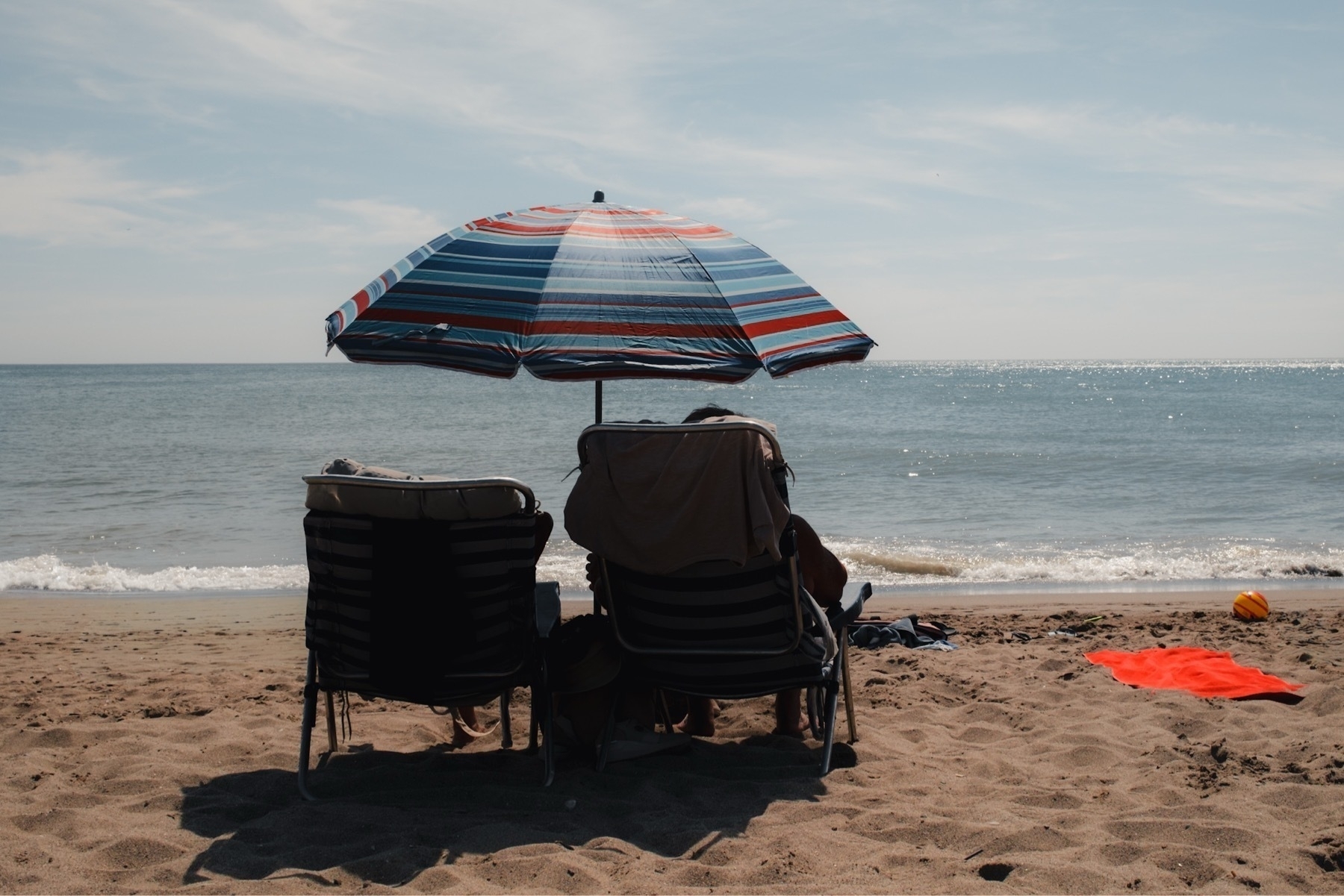 Two people sit under a striped umbrella on a sandy beach facing the ocean.