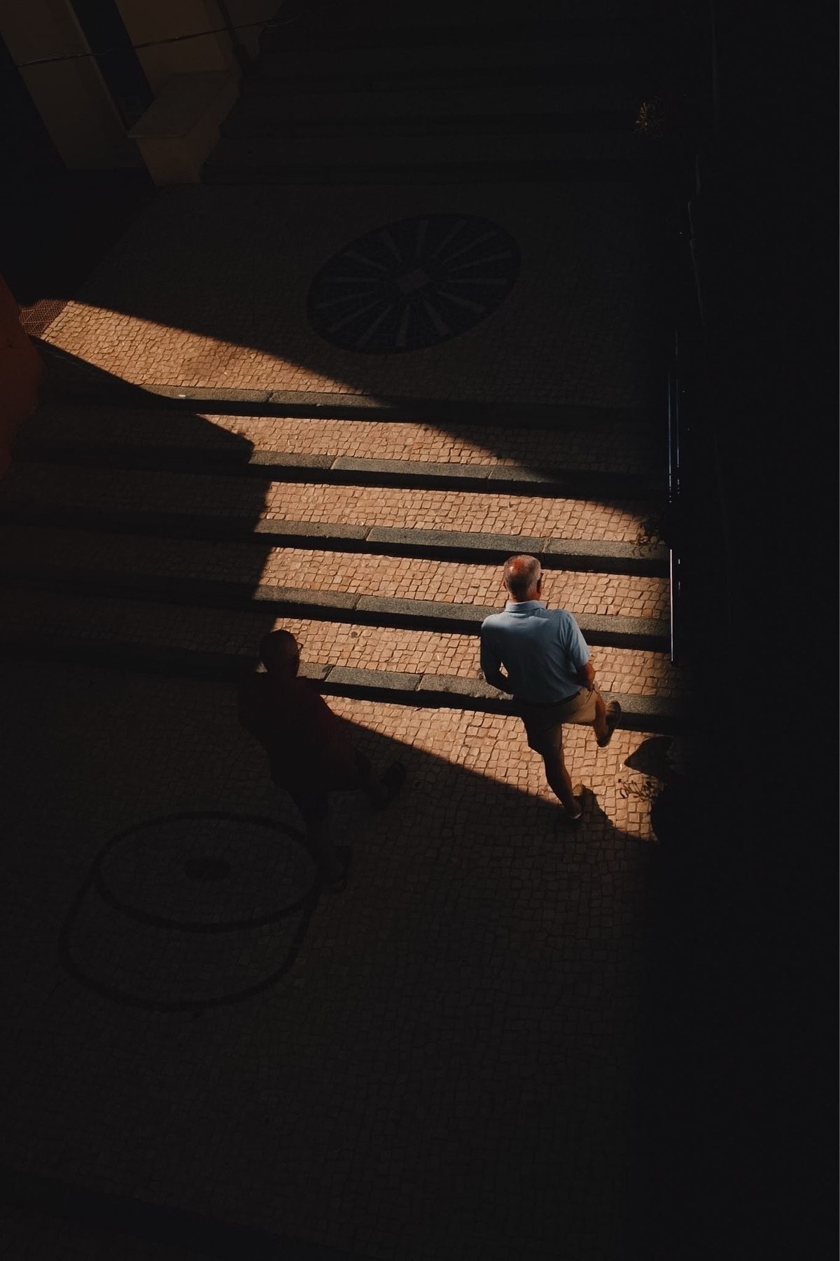 Two people are walking on a shadowed stairway, with sunlight illuminating part of the scene.