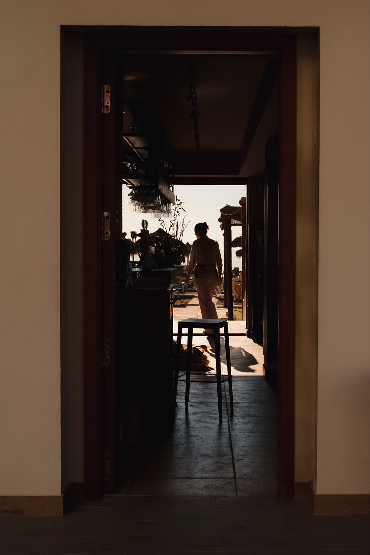 A person stands in a sunlit doorway, casting a shadow on the floor, with interior furnishings partially visible.