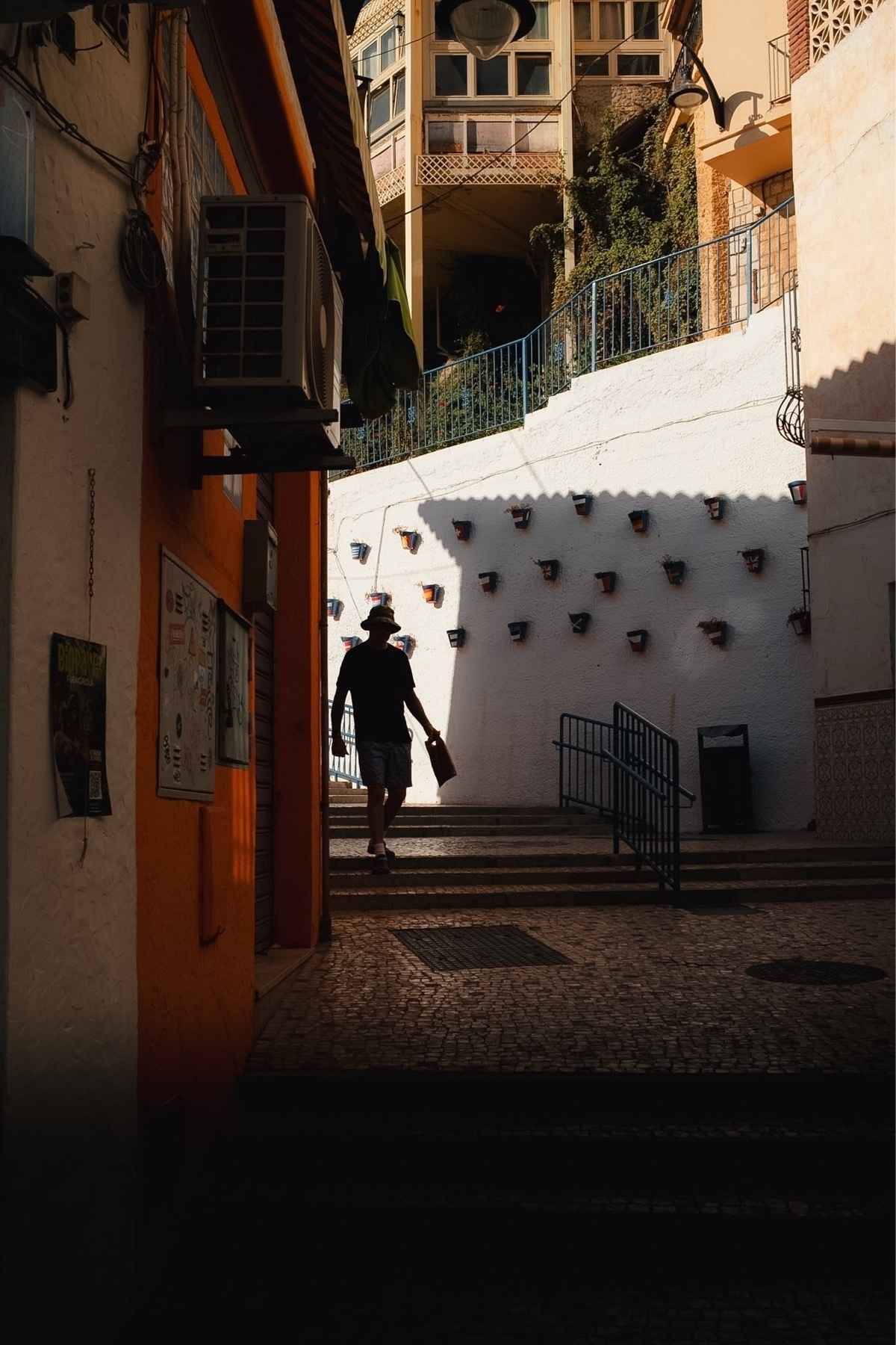 A person in a hat walks through a narrow, sunlit passage with a white wall adorned with potted plants and shaded areas.
