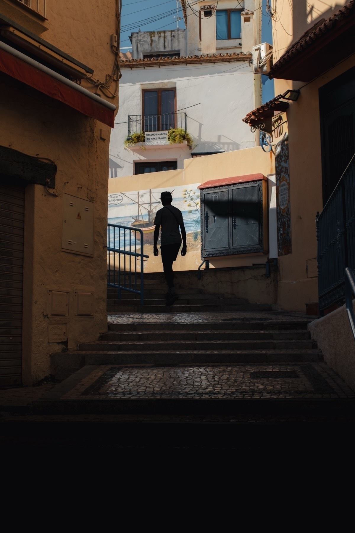 A person walks through a narrow, shadow-filled alley with sunlit buildings in the background.