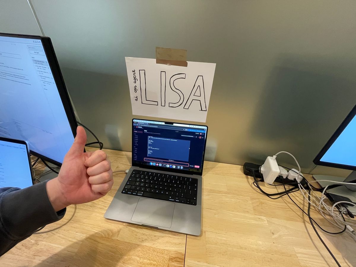 Photo of an AI agent which is the office's MacBook Pro with a thumbs up on the left and a name tag "Lisa' stuck on the office wall.