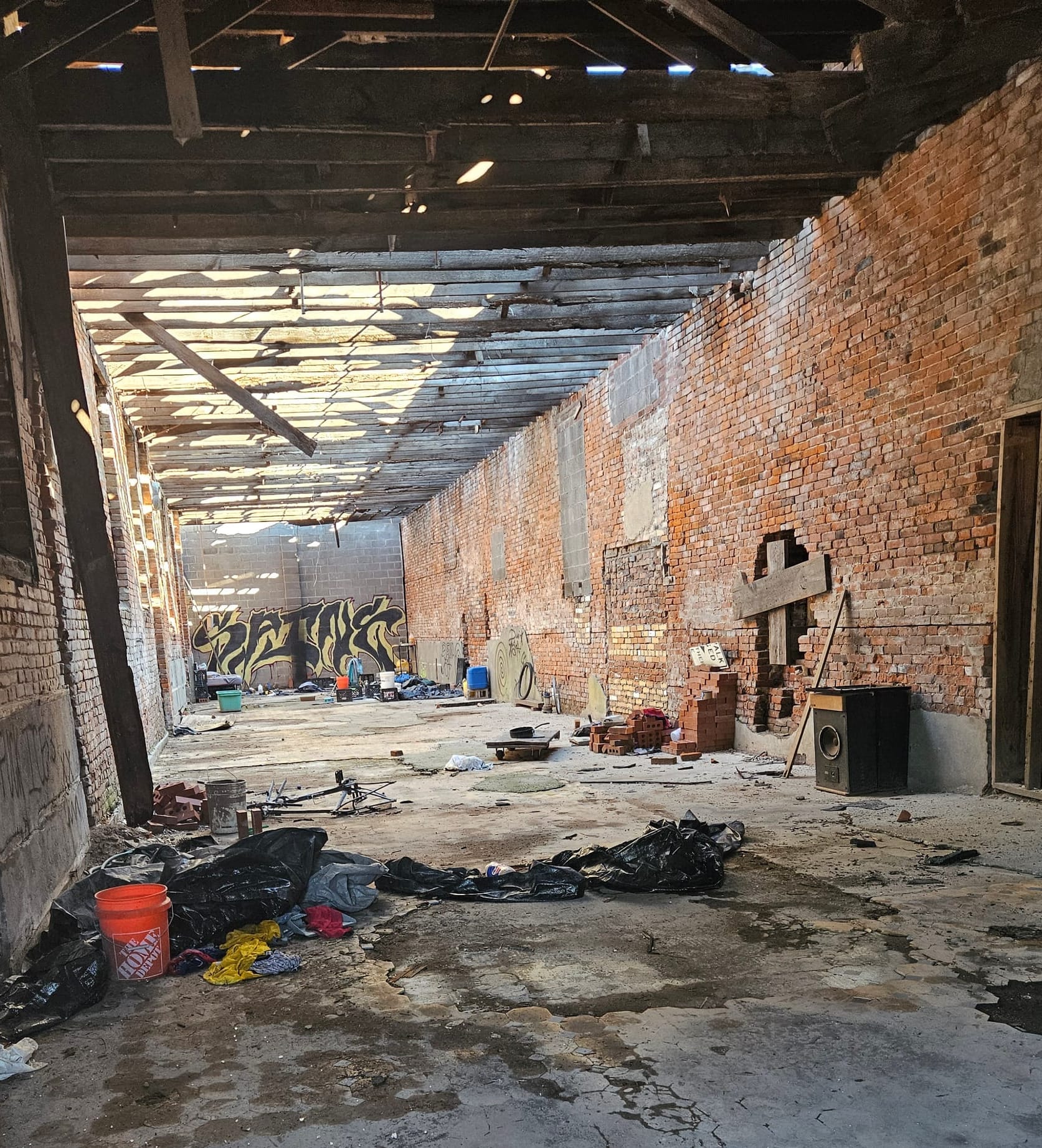 A worn out shell of a brick building. Trash lines the floor, and daylight is visible through the rafters. Graffiti highlights the rear wall. The visible wall is a cobbled patchwork of different colors of fill brick, wooden mending, and bricked-over windows.