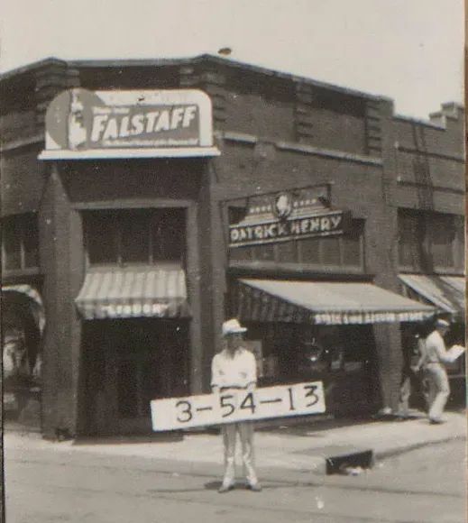 Black and white photograph of a corner building. The sign reads "Falstaff". A man stands in front of the building holding a placard with the tax ID number of the location.