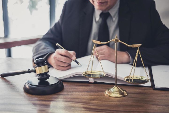 Advogado escrevendo em livro, com balança símbolo do direito em frente.