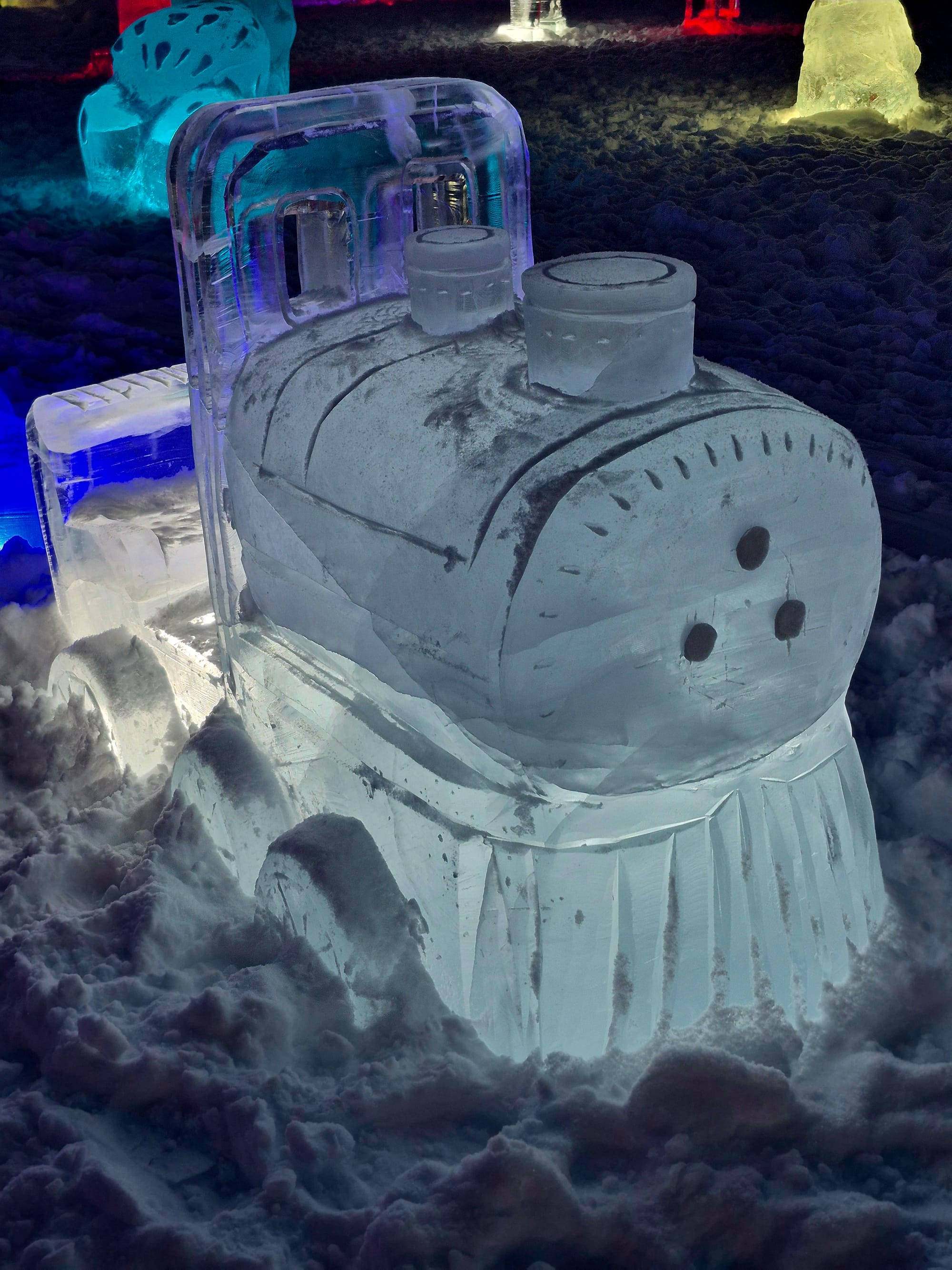 An ice locomotive glowing under colored lights — one of the public sculptures on display at the Fairbanks Ice Art Park alongside the championship works.