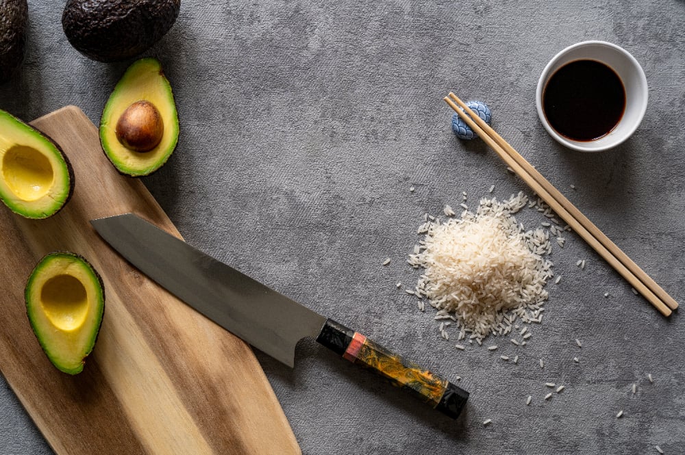 Top view of fresh avocados, a cutting board and knife, rice, and chopsticks on a grey surface
