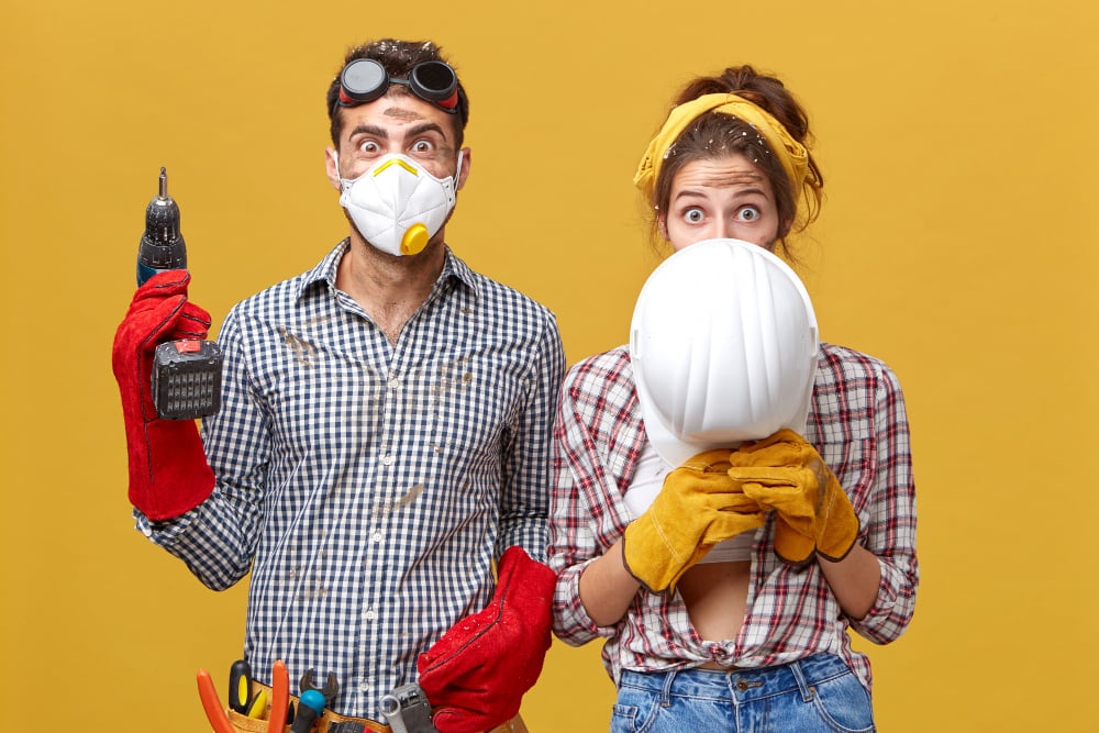 Male in protective mask standing with drill and female hiding under white hardhat renewing their flat working together using building instruments. Construction workers improving something in room