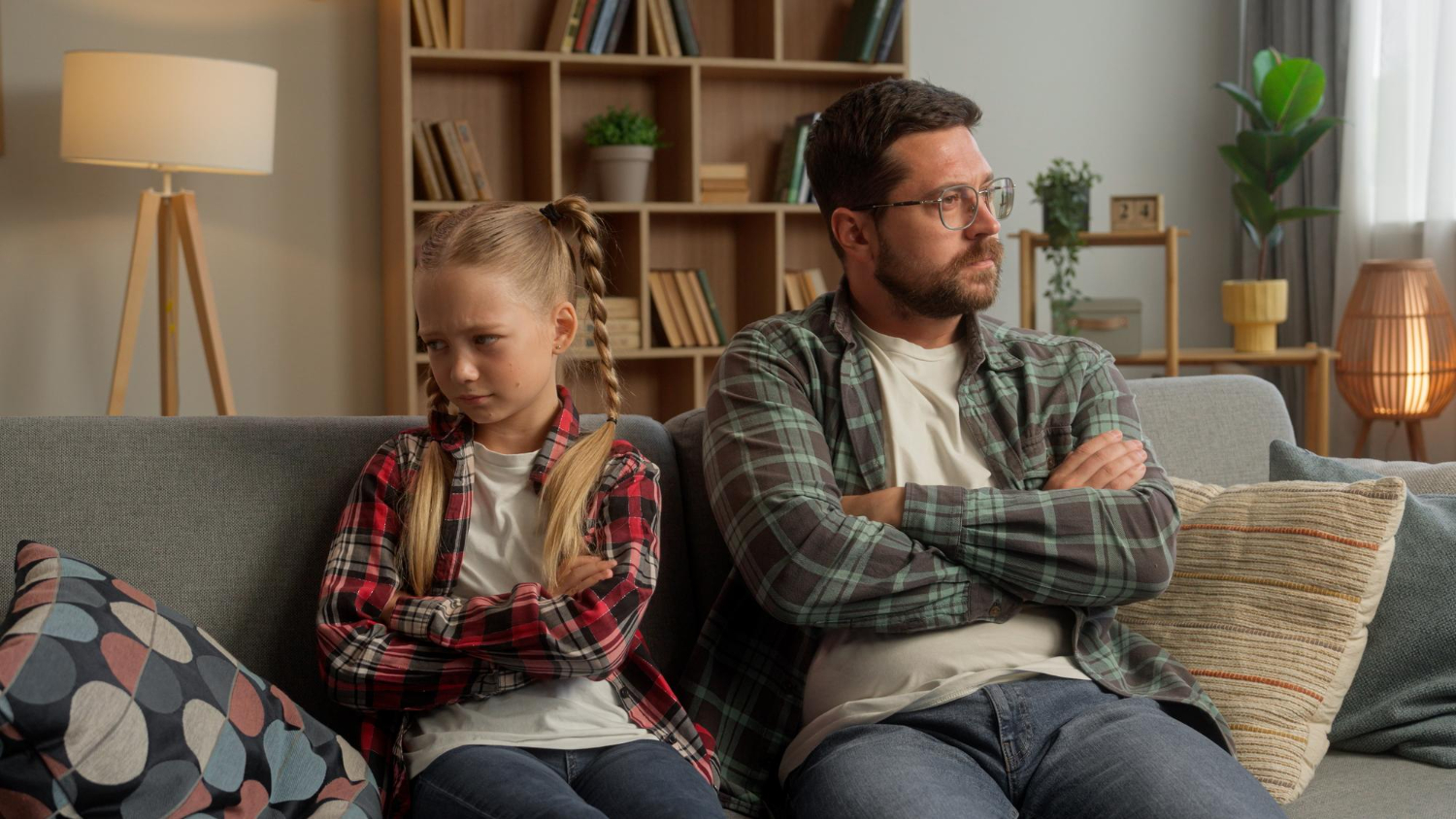 Frustrated dad and daughter sit with arms crossed on a sofa after a conflict—moment before choosing calm discipline.