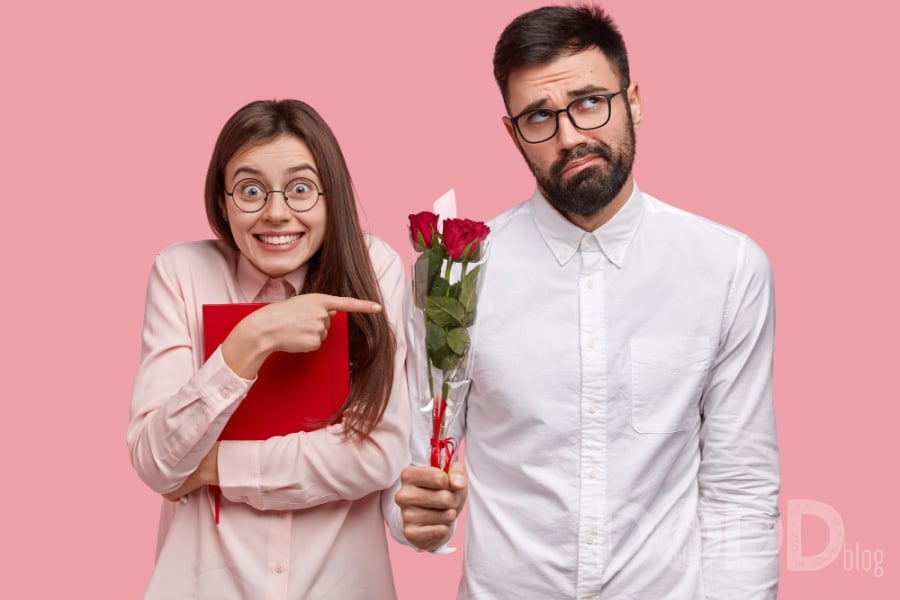 Positive young european woman carries red textbook, points at awkward bearded man in white shirt who feels shy, holds nice bouquet, have love story
