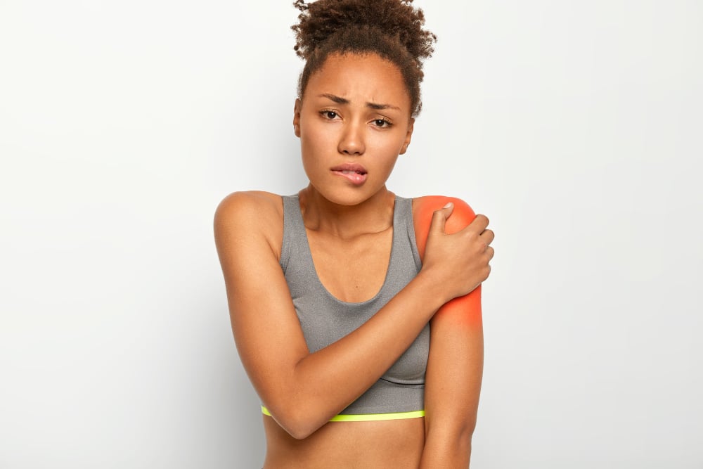 Free photo displeased curly haired woman bites lips, touches painful shoulder, suffers from injury, wears grey top, isolated on white background. health problems, medical concept