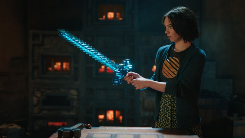 Cast of Minecraft The Movie - A girl stands confidently, holding a glowing, pixelated diamond sword inside a furnace-lit room. She’s focused and ready to fight.