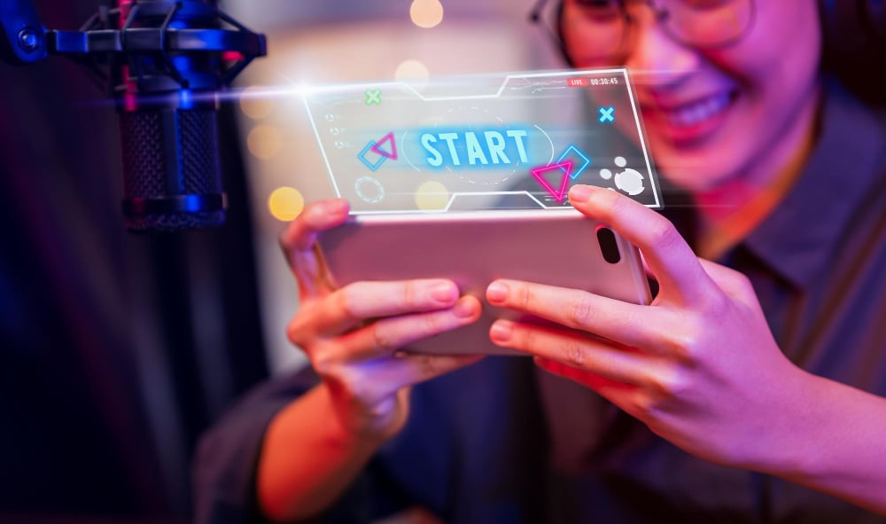 Excited young woman wearing headset and playing online game on smartphone with live broadcasting on internet
