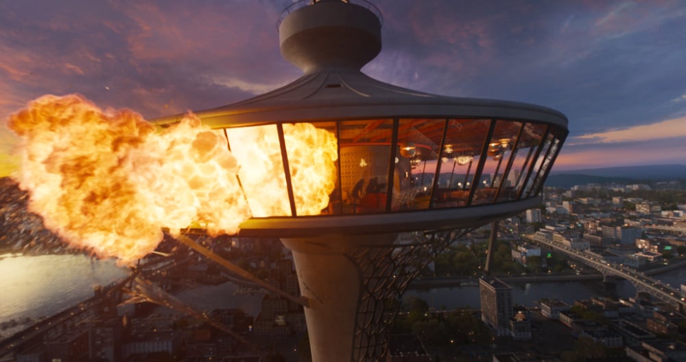A dramatic explosion engulfs a futuristic glass observation tower at sunset, high above a sprawling city skyline.