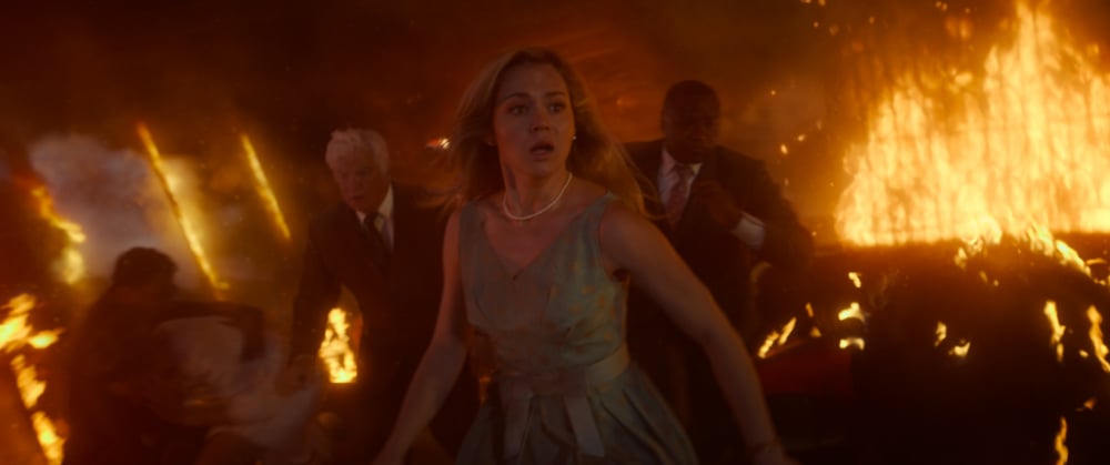 A woman in a sleeveless dress runs frantically through a burning building with flames erupting behind her, followed by other panicked individuals.