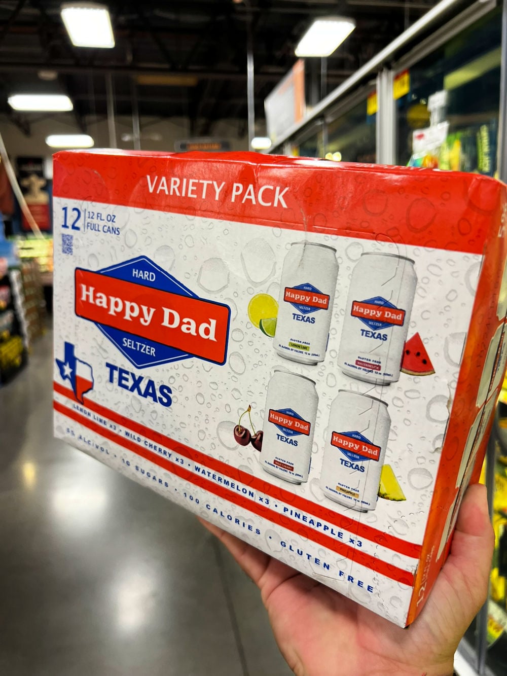 A person holding a 12-pack variety box of Happy Dad Texas Seltzer in a store aisle. The box displays four different flavors: Fruit Punch, Wild Cherry, Watermelon, and Pineapple. The Texas-themed packaging is prominently featured, with the box labeled 'Variety Pack' at the top.