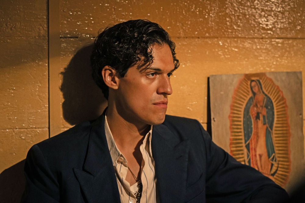 Henrique Zaga sits against a yellow wall with a solemn expression and a portrait of the Virgin of Guadalupe behind him.