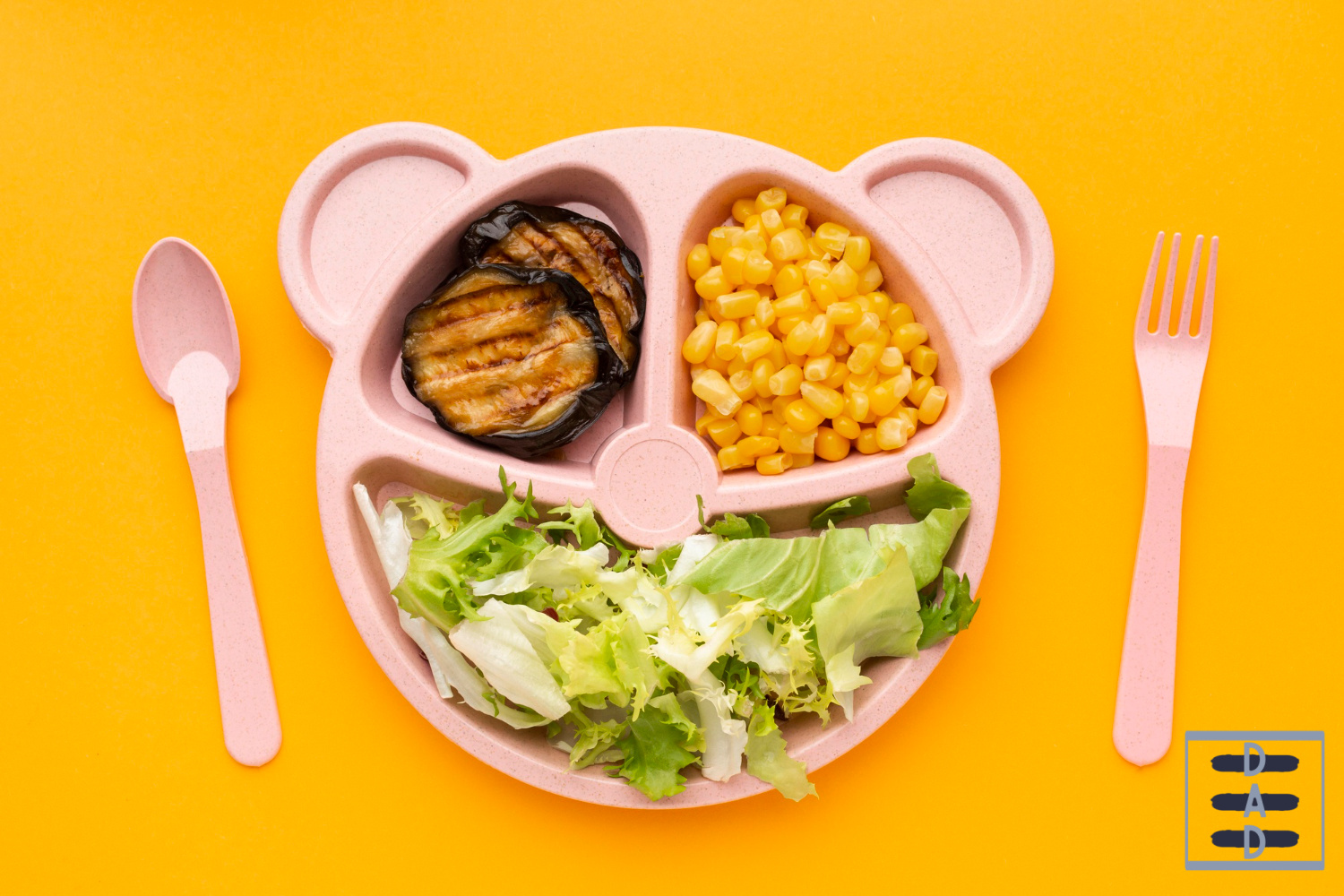 A pink bear-shaped kids lunch plate with grilled eggplant, corn, and fresh salad on a bright yellow background with pink fork and spoon.