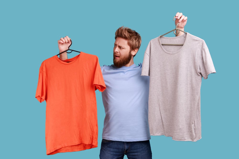 Man frowning face holding two hangers with gray and orange Recycled T-Shirts hard to choose the best attire.
