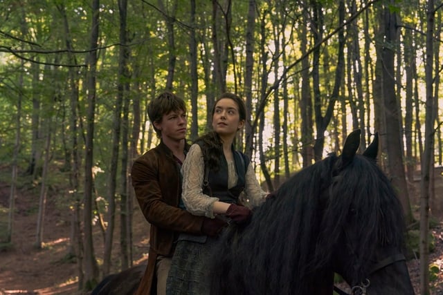 A young man and woman riding a black horse through a dense, green forest. The man looks protective, while the woman appears determined, gazing ahead.