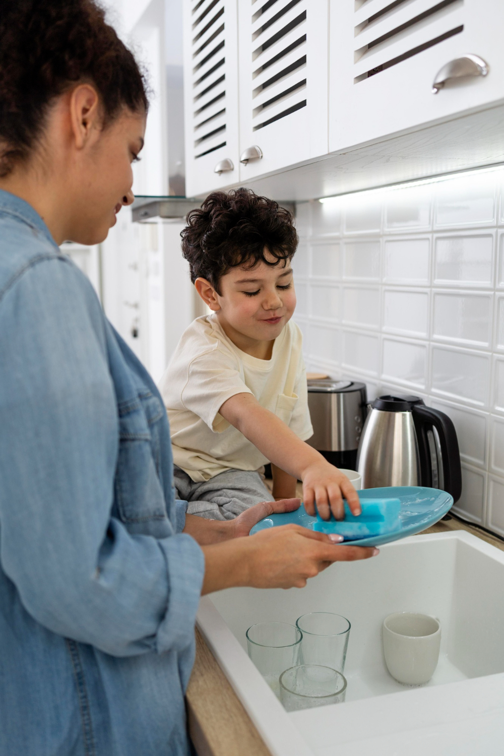 Mother and young son washing dishes together in the kitchen, turning chores into fun.
