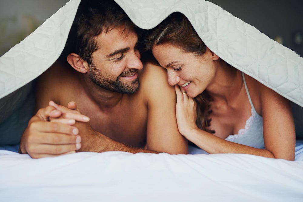 Romantic couple smiling and cuddling under a blanket on a bed, sharing an intimate and joyful moment.
