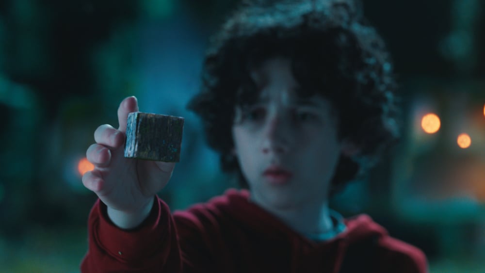A young boy with curly hair, dressed in a red hoodie, holds up a mysterious block, gazing at it with curiosity and focus.