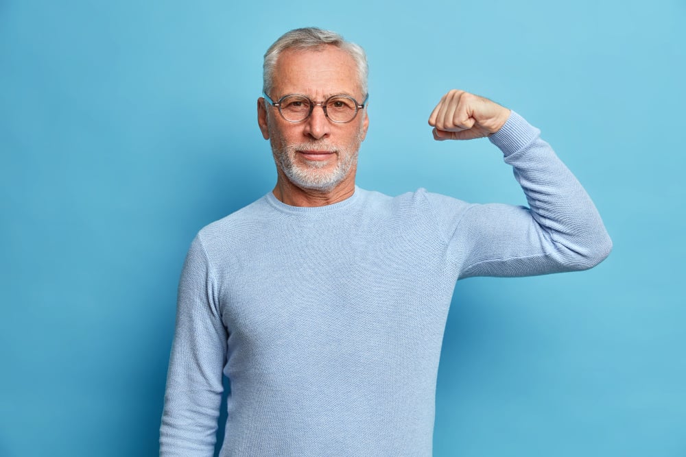 Senior bearded man shows muscles after practising bodybuilding wears transparent glasses and basic jumper poses against blue studio wall
