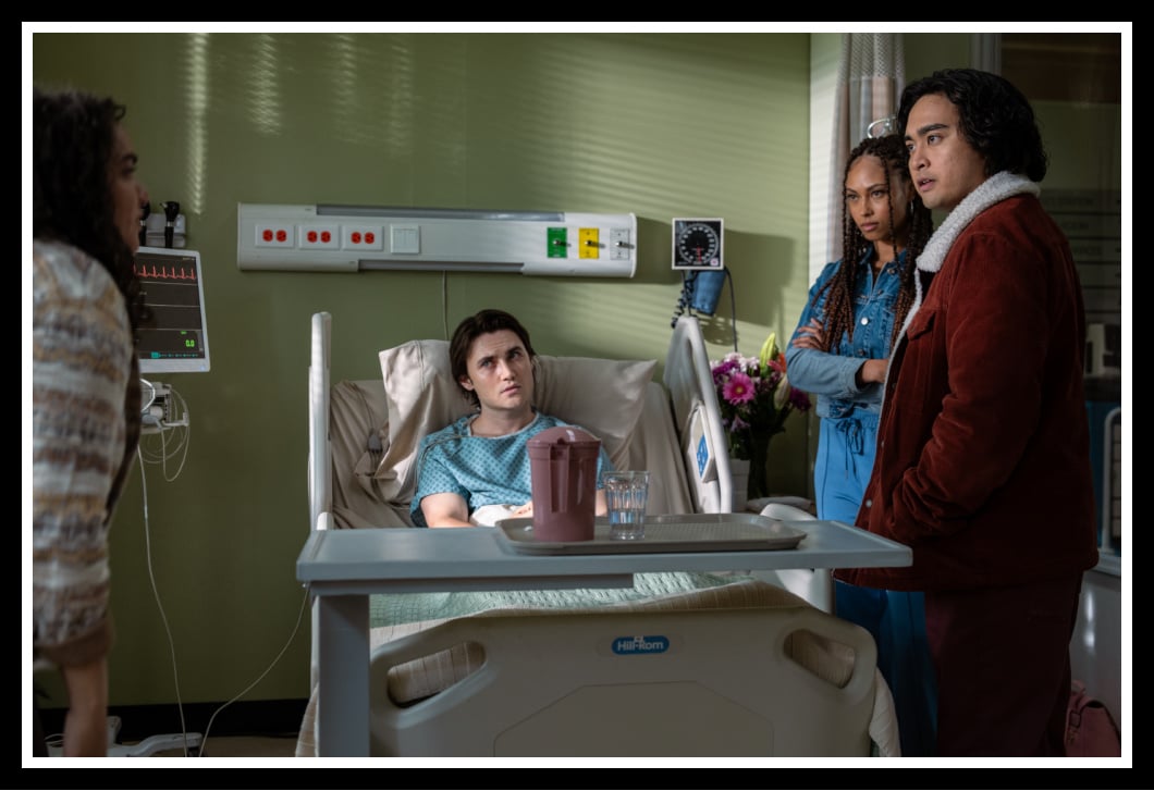 Simon Elroy (Kristian Ventura) lies in a hospital bed, surrounded by Nicole Herrera (Kiara Pichardo) and other concerned friends in a tense moment from School Spirits Season 2.