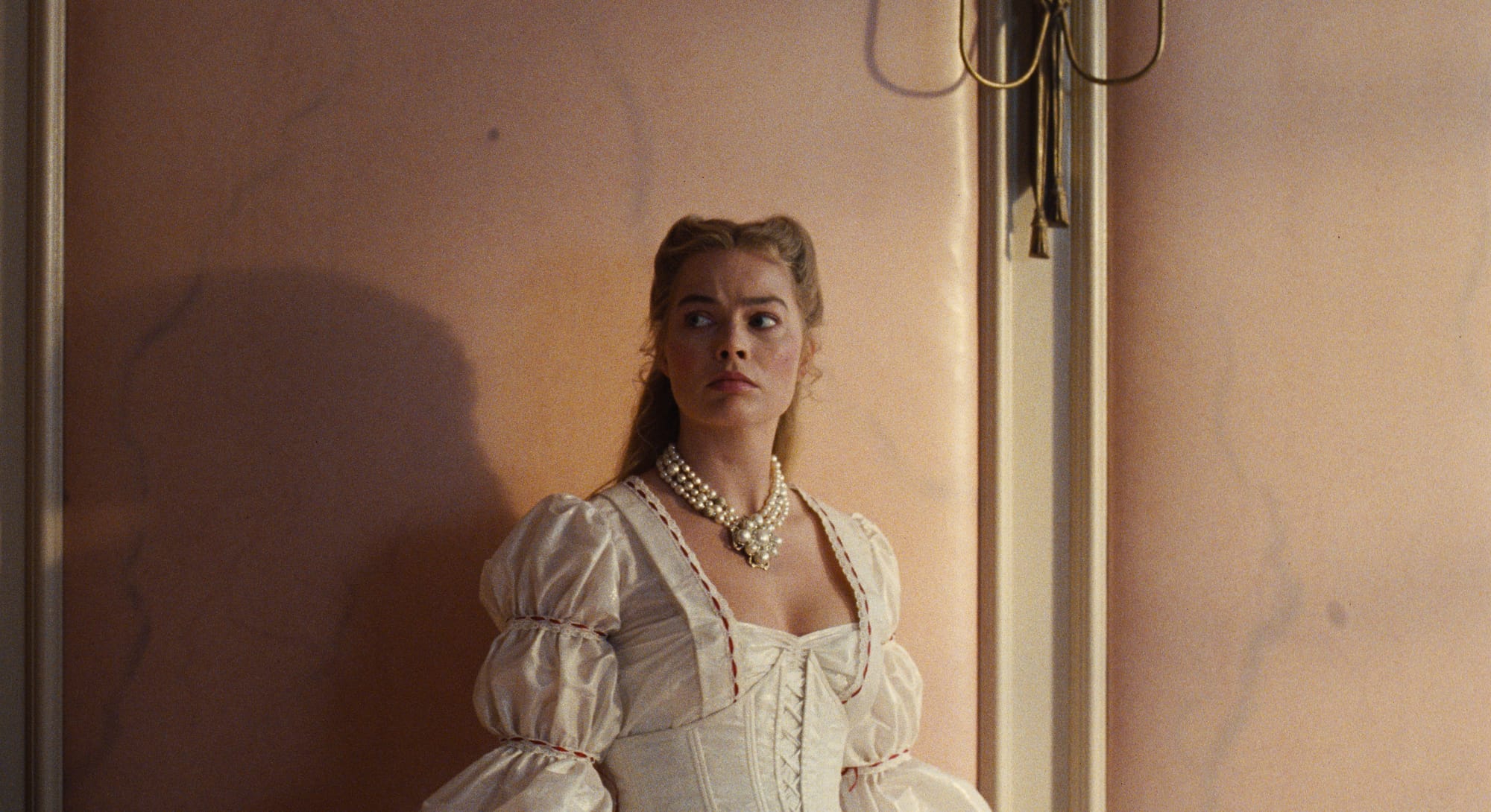 Margot Robbie as Catherine stands against a wall in a tense scene from Wuthering Heights (2026), reflecting emotional distress, psychological punishment, and unhealthy relationship dynamics.