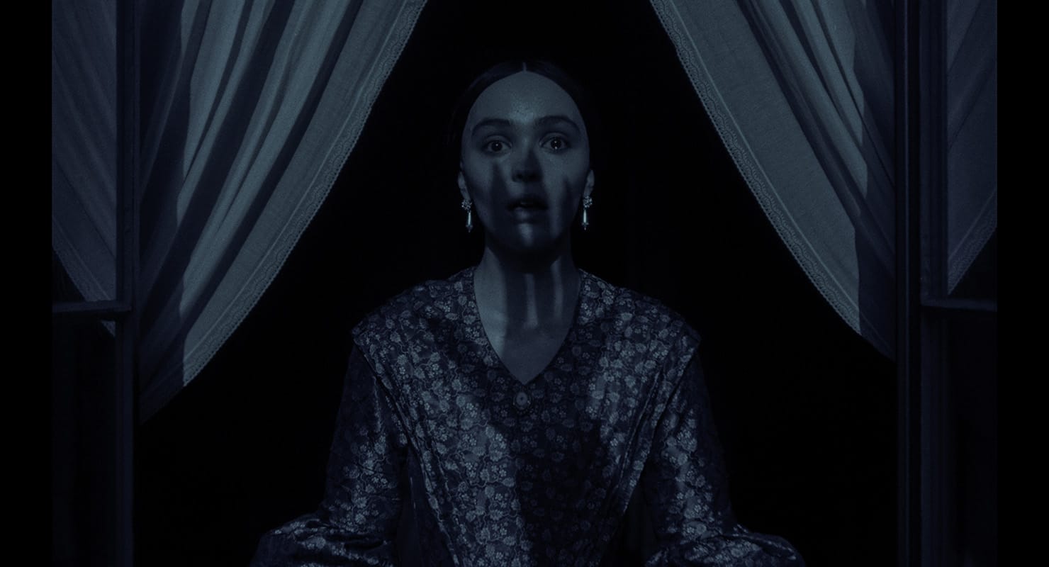 A haunting cinematic shot from Nosferatu (2024) showing Ellen Hutter (Lily-Rose Depp) standing at a window in a state of distress and alarm.