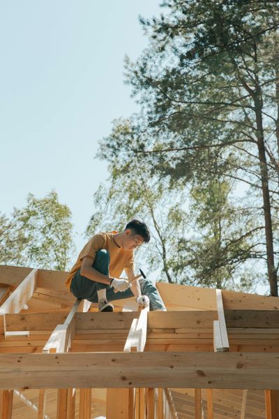 A carpenter works on a wooden roof frame outdoors under clear skies.