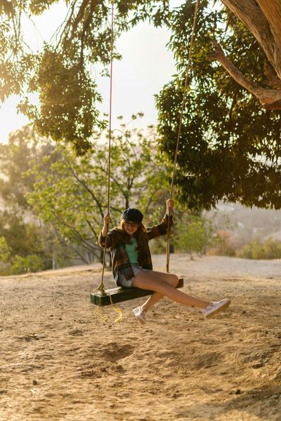 A happy young girl swings on a rope swing hanging from a tree in a sunny outdoor setting.