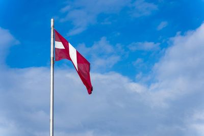A Latvian flag on a flagpole waving against a backdrop of clear blue sky and clouds.