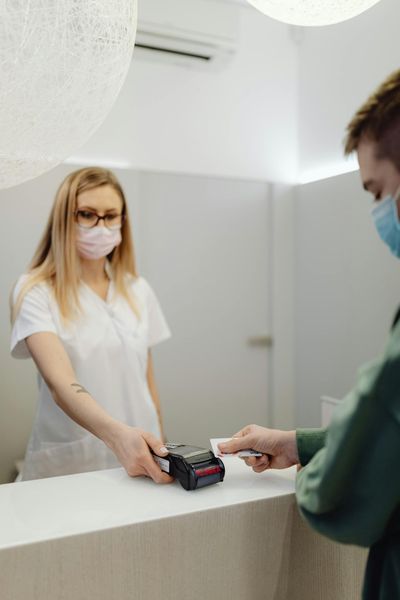A masked patient pays at a clinic counter, using a card terminal handled by a healthcare professional.