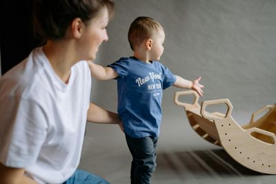 A young boy in a blue shirt playing with a wooden rocking toy under adult supervision indoors.